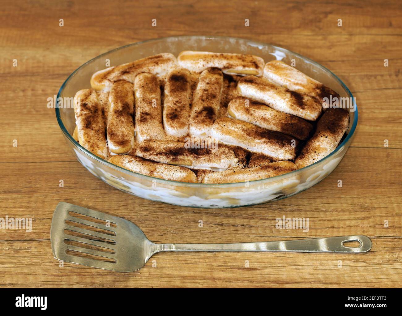 Tiramisu mit Kaffee getränkten Marienfinger und Mascarponcreme, mit Kakao in einer ovalen Glasschale auf einem Holztisch bestäubt, Metallspachtel an der Seite, Re Stockfoto
