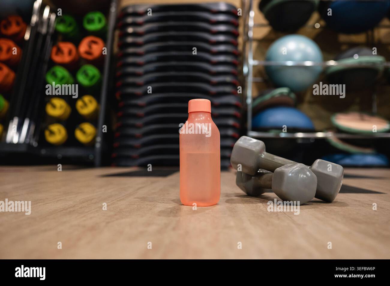 Wiederverwendbare Wasserflasche mit Kurzhanteln auf dem Boden des Fitnessstudios. Fitnessgeräte im modernen Sportclub-Innenraum. Stockfoto