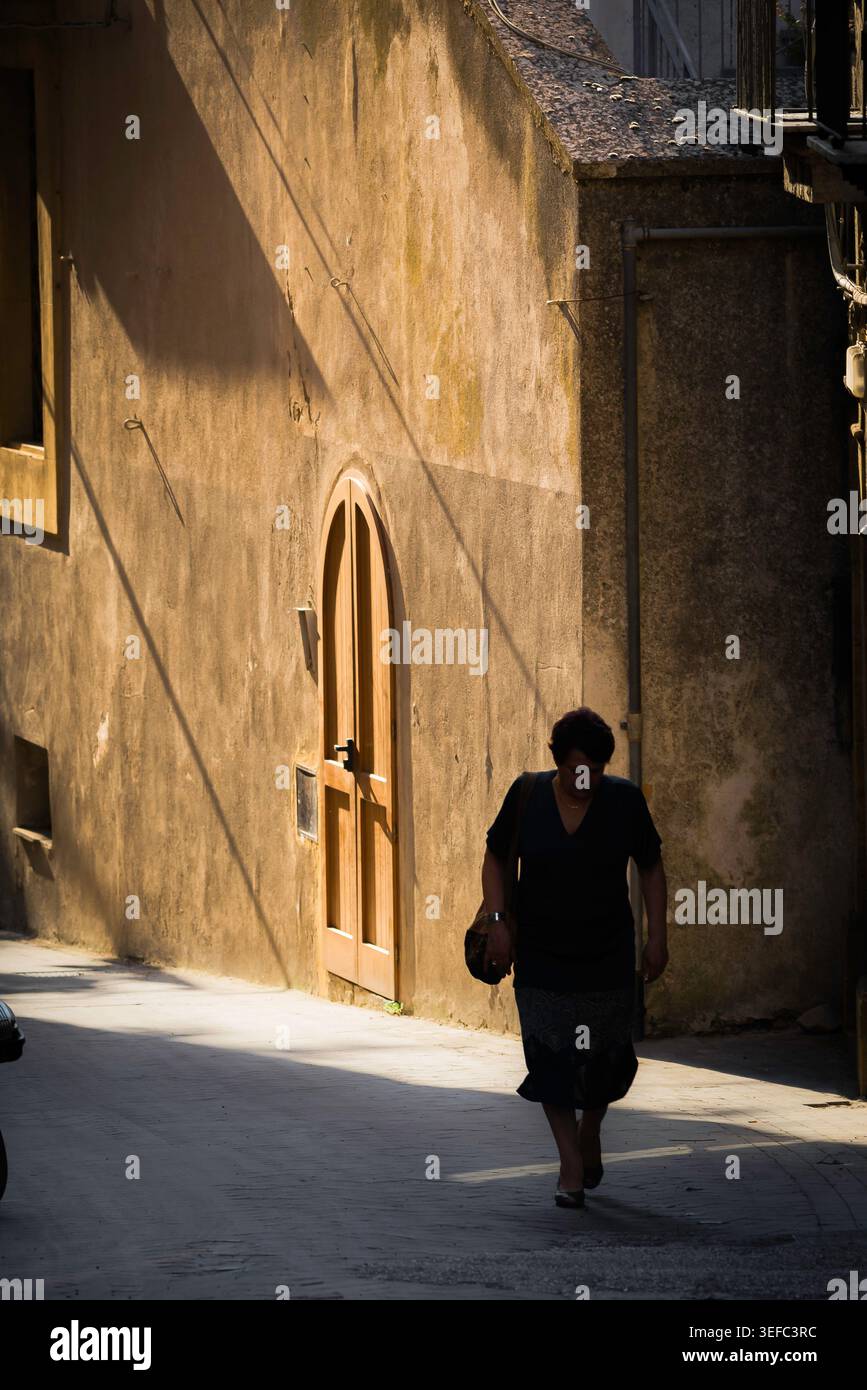 Einsame Frau, Blick auf eine Frau mittleren Alters, die allein im Schatten in einer leeren Straße in der historischen Altstadt von Enna, Sizilien, spaziert. Stockfoto