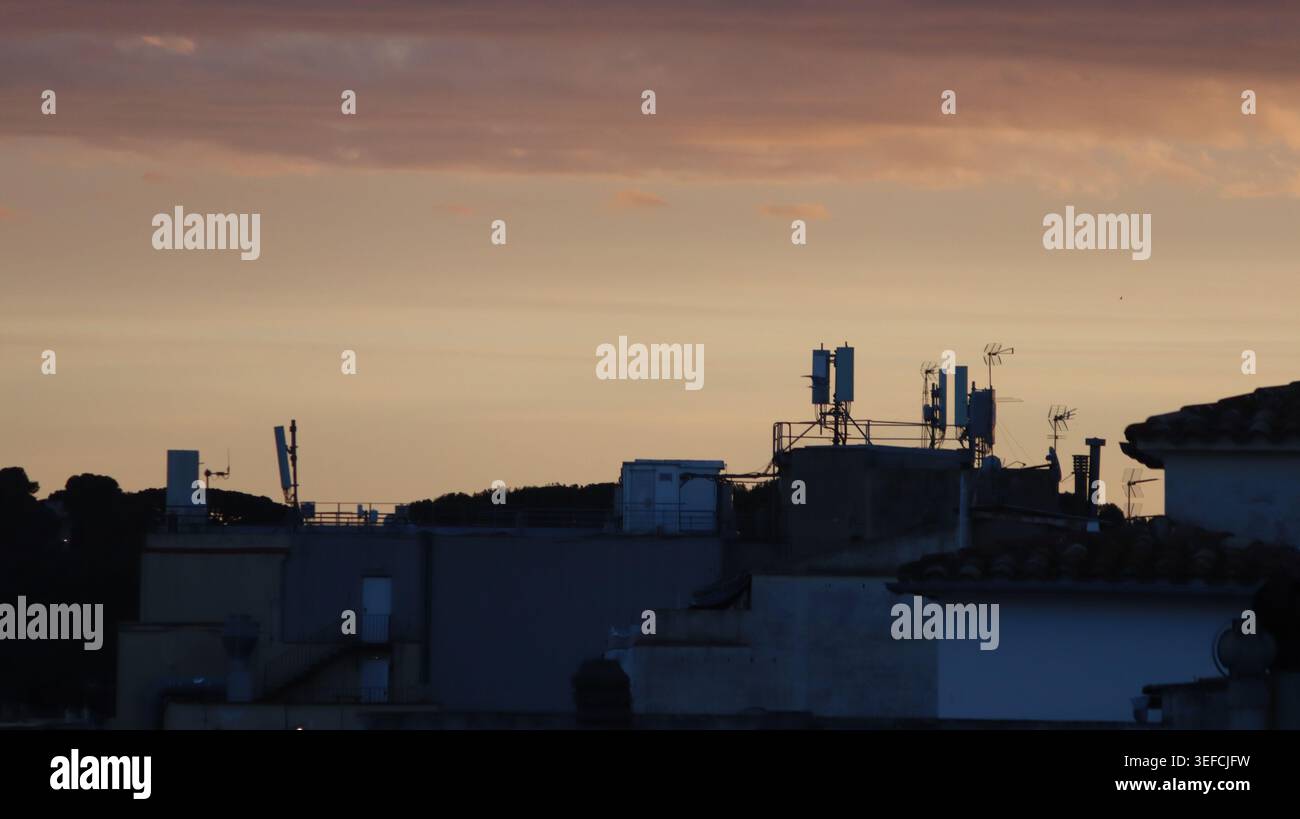 Dächer von Stadtgebäuden mit Mobilfunkantennen und Kommunikationstürmen, die vor einem warmen, orangefarbenen Himmel mit Sonnenuntergang stehen. Stockfoto