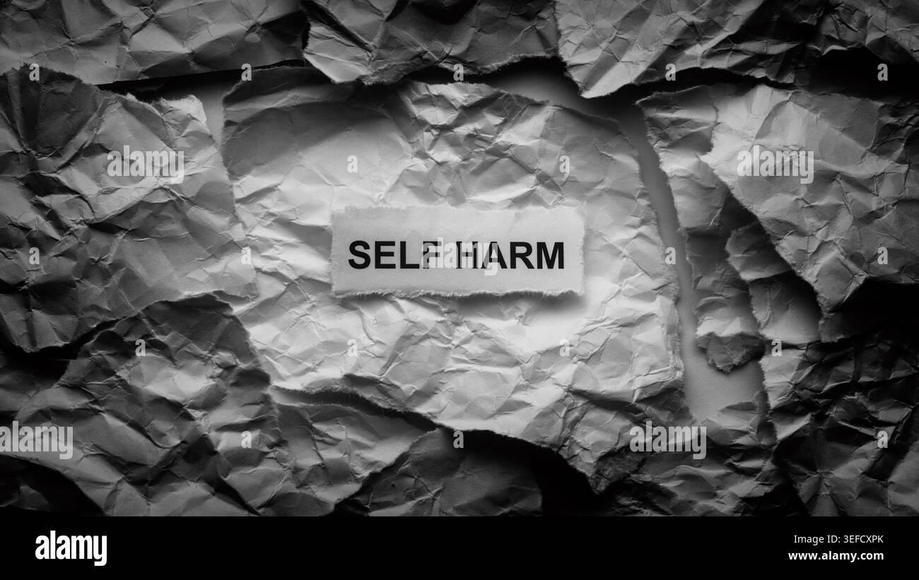 Ein zerrissenes Stück Papier mit den Worten Self Harm auf einem Hintergrund aus chaotischen zerknitterten Papierstrukturen. Schwarz-weiß. Nahaufnahme. Stockfoto