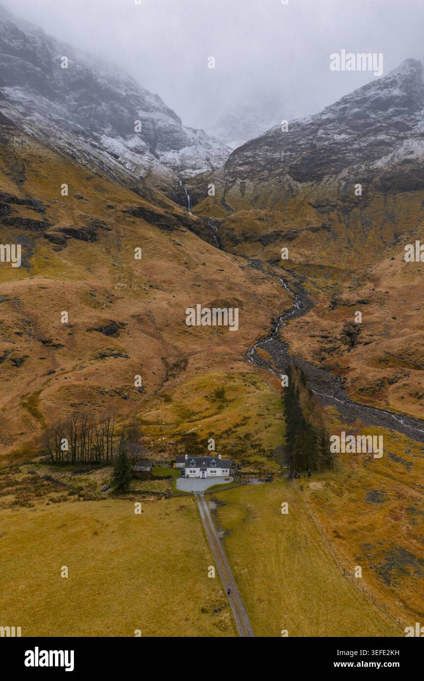 Blick aus der Vogelperspektive auf das berühmte weiße Cottage am Fuße der schneebedeckten Berge mit einem gewundenen Bach und zerklüftetem Gelände Glencoe, Schottland, Unite Stockfoto
