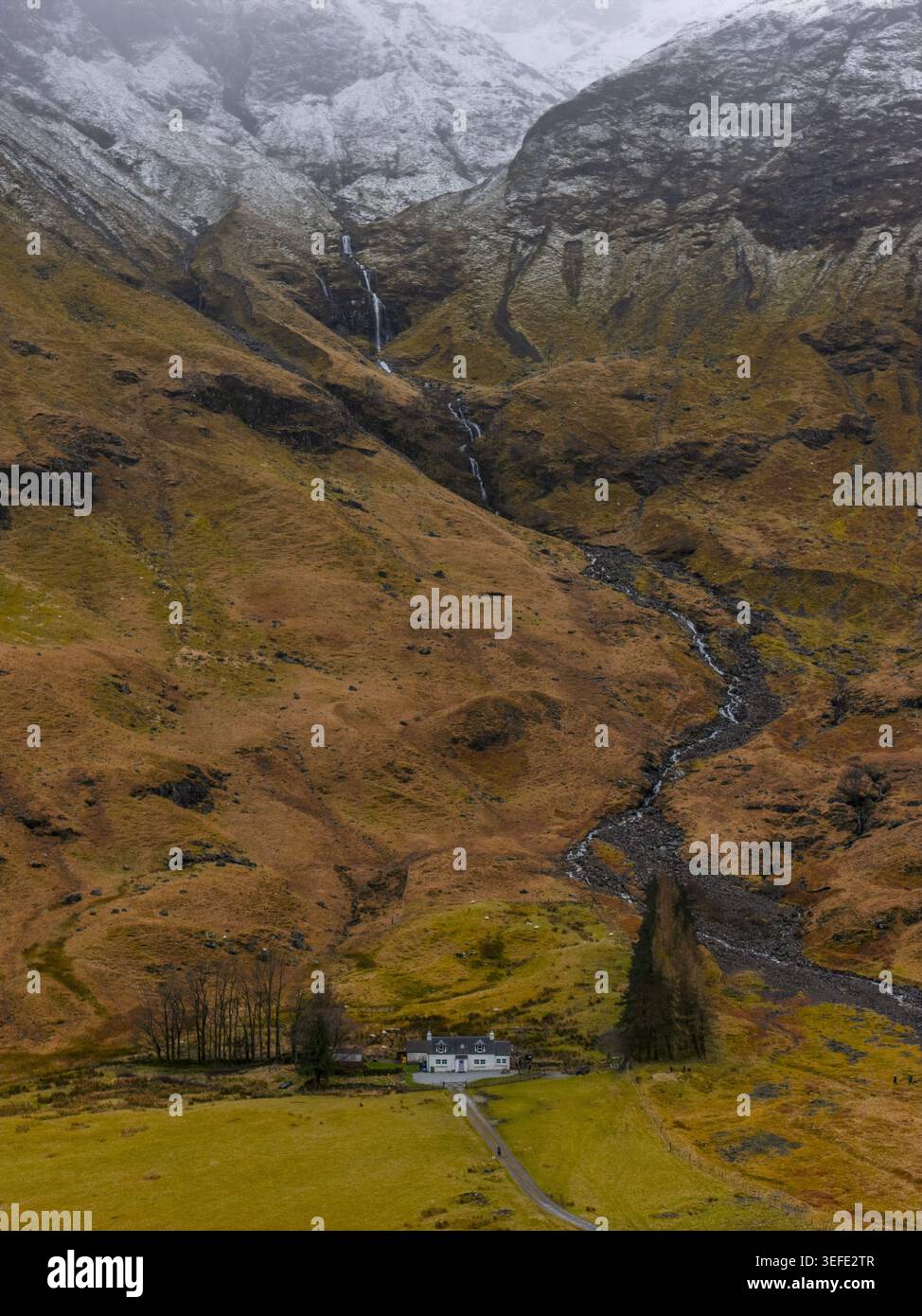 Aus der Vogelperspektive eines einsamen weißen Cottage am Fuße der zerklüfteten Berge mit einem Wasserfall und schneebedeckten Gipfeln Glencoe, Schottland, Vereinigtes Königreich Stockfoto