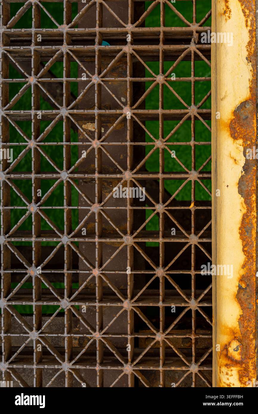 Das Metallgitter zeigt ein Muster aus Dreiecken und Quadraten mit Rostflecken. Der Hintergrund ist ein lebhaftes Grün, das auf eine städtische Umgebung hindeutet. Diese sc Stockfoto