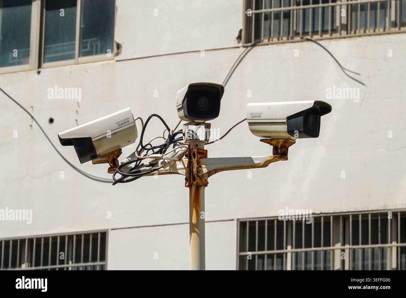 HAINING, CHINA - 1. MAI 2026: Drei Hikvision-Sicherheitskameras, die auf einem rostigen Pfosten gegen ein Gebäude mit vergitterten Fenstern montiert sind, symbolisieren den Omnip Stockfoto