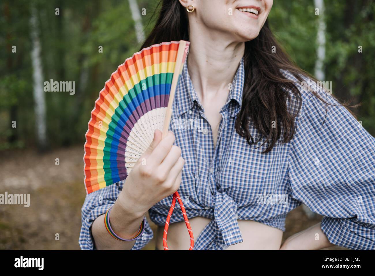 Die Person lächelt, während sie den Regenbogenventilator im Freien hält. lgbtq-Identitätsdarstellung, queerer persönlicher Stil, Geschlechterausdruck. Stockfoto
