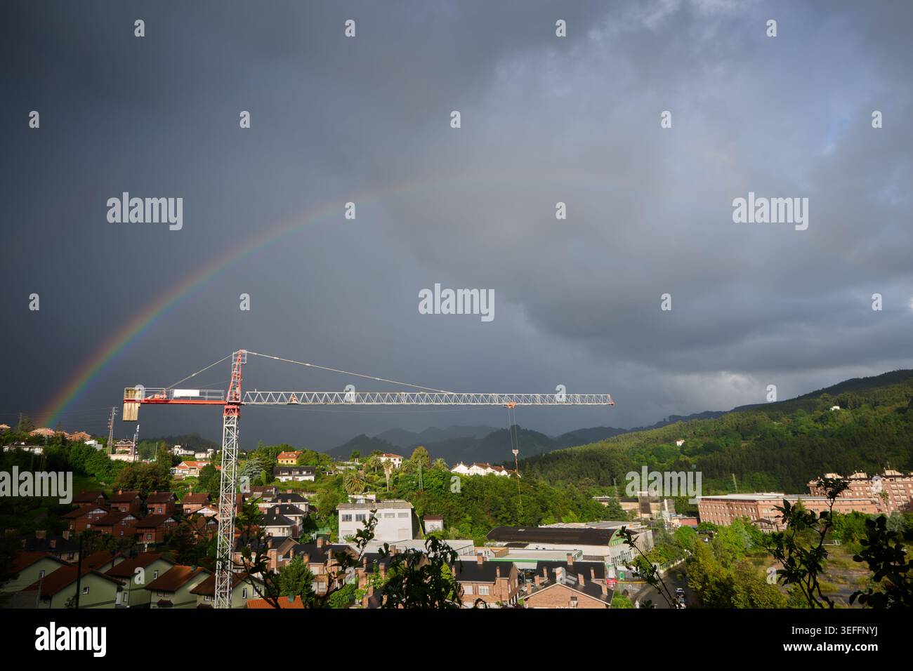 Heller Regenbogen, sichtbar über einem Baukran und Wohngebiet, mit dunklen Sturmwolken an einem anspruchsvollen Tag Stockfoto