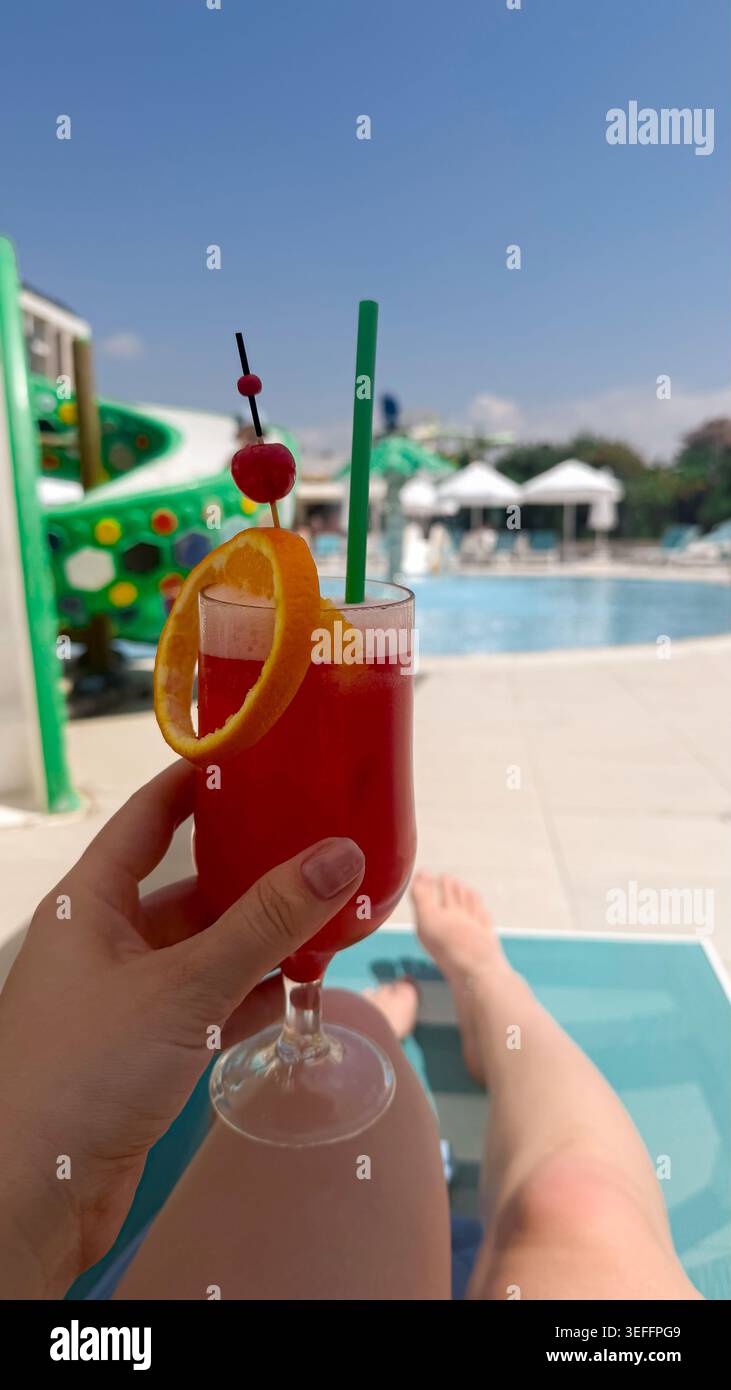 Halten Sie einen farbenfrohen Cocktail mit Orangenscheiben und Kirsche, Blick auf den Pool mit Wasserrutsche und Liegestühlen in einem Resort-Ambiente Stockfoto