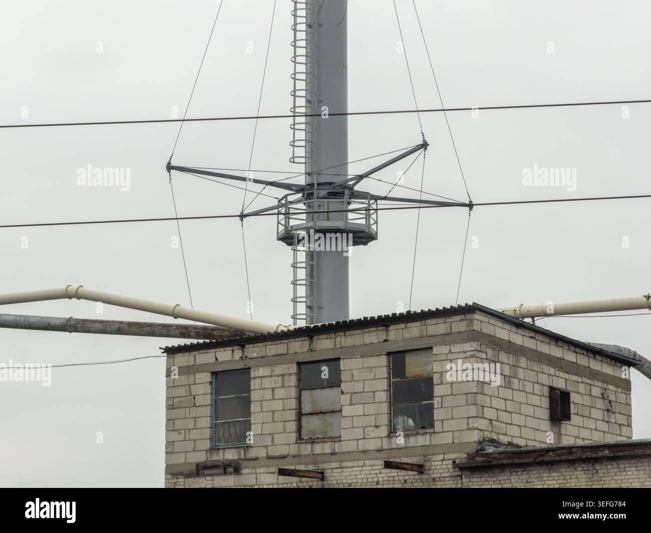 Funkmast aus Metall mit Dienstplattform und Kabeln, die über einer industriellen Struktur aus Ziegelsteinen ragen und Telekommunikationsnetze und Stromverteiler hervorheben Stockfoto