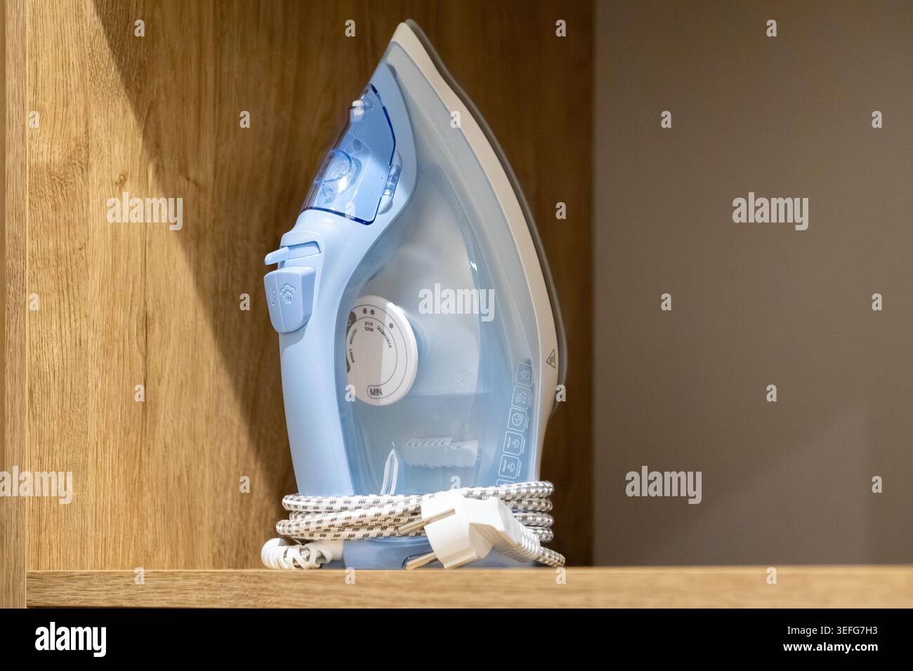 Nahaufnahme eines blauen elektrischen Dampfbügeleisens, das aufrecht auf einem Regal mit umwickeltem Netzkabel steht und die Bekleidungspflege, die Haushaltswartung und das Interio-System veranschaulicht Stockfoto