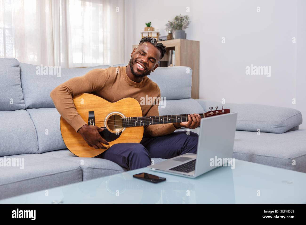 Eine Person sitzt bequem auf einer Couch und spielt eine Akustikgitarre, während sie Online-Gitarrenunterricht nimmt. Die entspannte Atmosphäre wird durch n Stockfoto