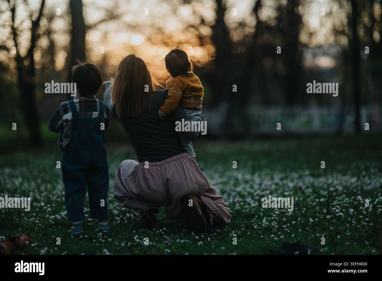Mutter kniet mit zwei Kindern in einem Park bei Sonnenuntergang, Familienmoment draußen Stockfoto