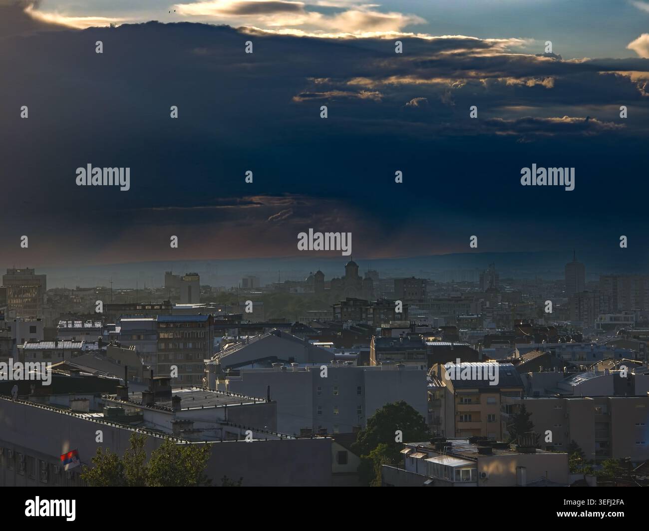Sonnenstrahlen brechen durch schwere Sturmwolken über Belgrad, Serbien, und beleuchten Dächer, Kirchtuppeln, ferne Hügel und eine serbische Flagge in einem dramatischen E Stockfoto
