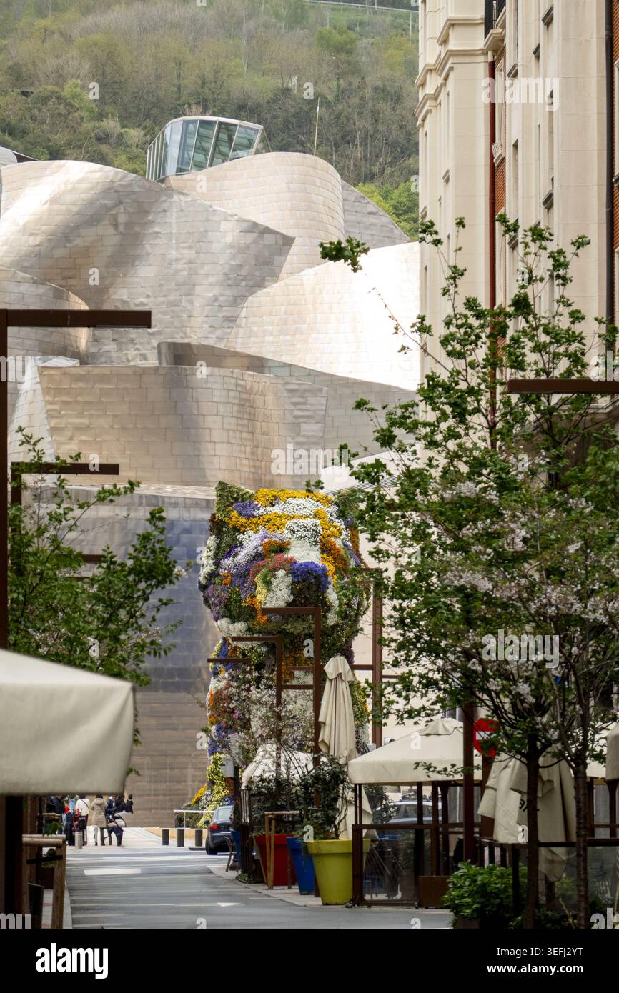 Blick auf das Guggenheim Museum Bilbao mit seinen Titankurven und die Blumenskulptur des Hündchens aus einer Straße mit Bäumen und Gebäuden in Bilbao, EU Stockfoto