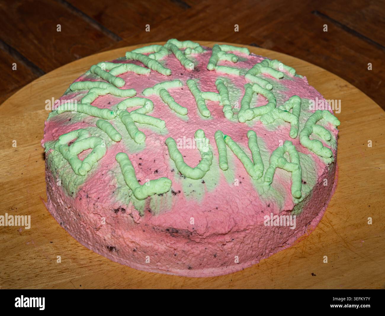 Hausgemachte rosa Milchkuchen mit grünen Zuckerglasbuchstaben, die alles Gute zum Geburtstag Luna auf Holzbrett für Kinderparty mit rustikalem, handgemachtem Charme buchstabieren. Stockfoto