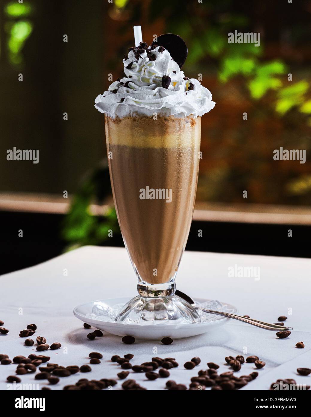 Kaffee-Milchshake mit Schlagsahne und Schokoladenspänen. Kakao wirbelt in hohem Glas, serviert auf weißer Untertasse. Kaffeebohnen, die um b verteilt sind Stockfoto