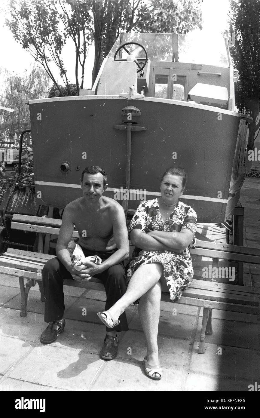 Sedove, 1982, ruht vor einem berittenen Boot. Ein Vintage-Schwarz-weiß-Foto mit einem entspannten verheirateten Paar im Urlaub. Kravelis Michail Iljitsch und Kravelis Valentina Timofeevna sitzen auf einer Holzbank, Valentina kreuzt ihre Beine und faltet ihre Arme bequem. Sie sitzen im Schatten eines großen Bootes, das als Parkdekoration dargestellt wird, und genießen das ruhige Ambiente des Badeortes. Das Foto verkörpert die Atmosphäre einer vergangenen Ära und die Erinnerung an das Leben vor dem Krieg. Stockfoto