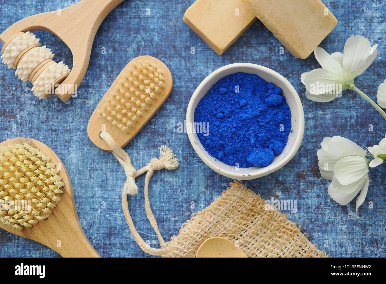 Natürliche Spa-Bürsten, blaues Pigmentpulver, Seife und Blumen auf rustikalem blauem Stoffhintergrund Stockfoto