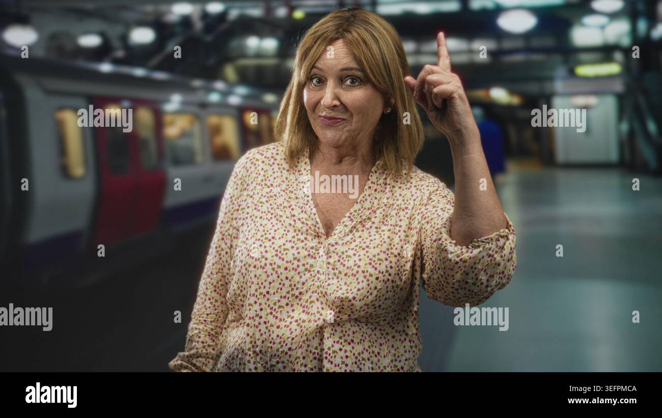 Die Frau zeigt den Zeigefinger nach oben in ein U-Bahn-Bahnsteig-Gebäude, lächelt in einer Blumenbluse und sichtbarem Unterarm; Neugier. Stockfoto