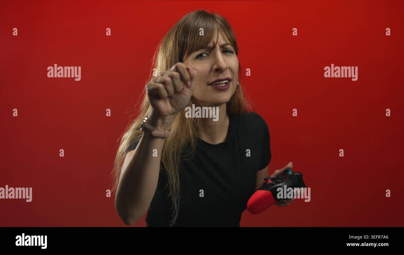 Junge Frau mit langen Haaren, die einen roten Spielcontroller hält, die Faust hochgezogen und im Studio vor rotem Hintergrund grimmig; Frustration. Stockfoto