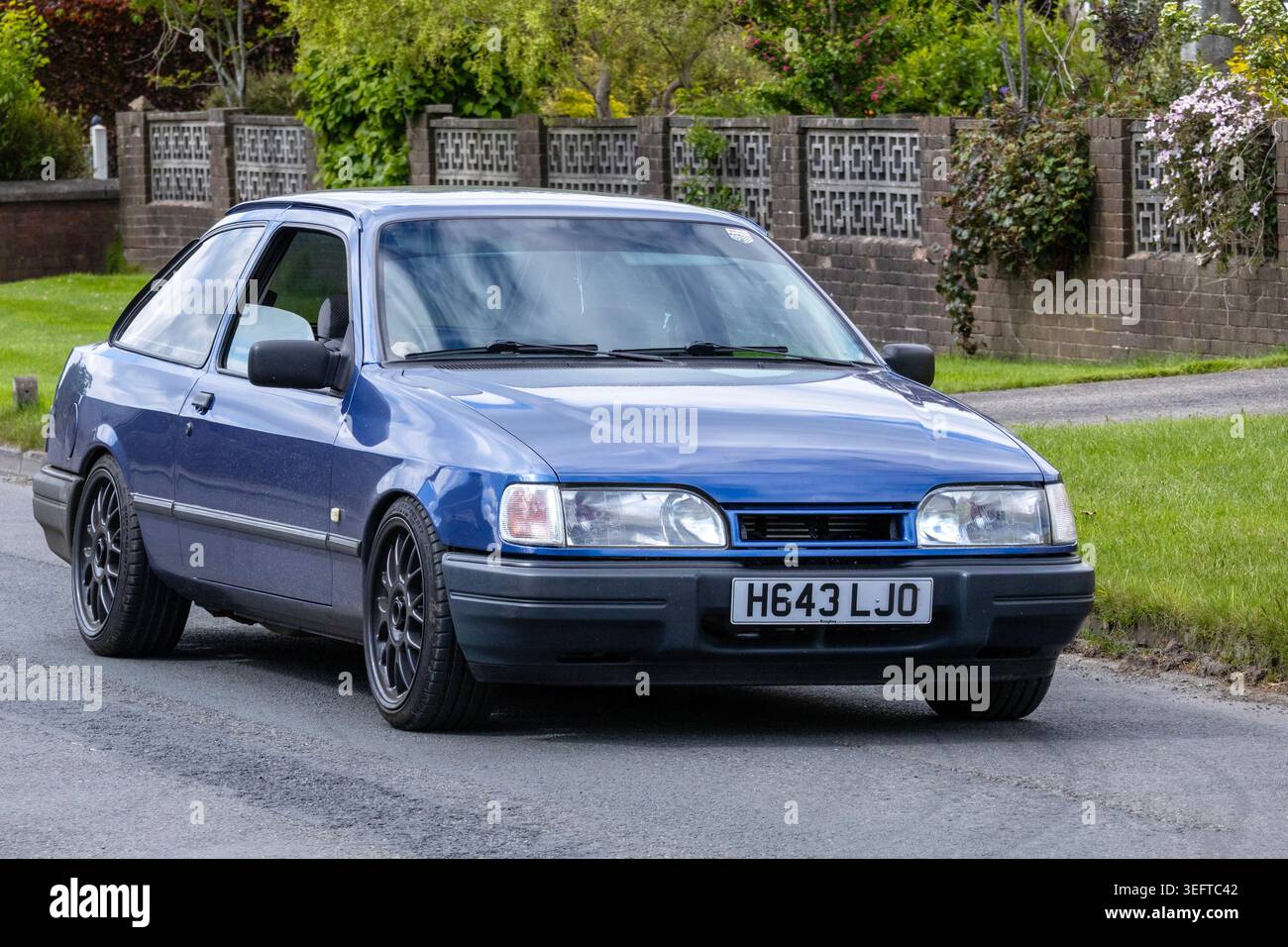 Ein klassischer, blauer Ford Sierra 1990 dreitüriger Fließheck mit speziell angefertigten Leichtmetallrädern fährt auf dem Weg zur Renntag Car Show 2026 auf einer Wohnstraße in Großbritannien. Stockfoto