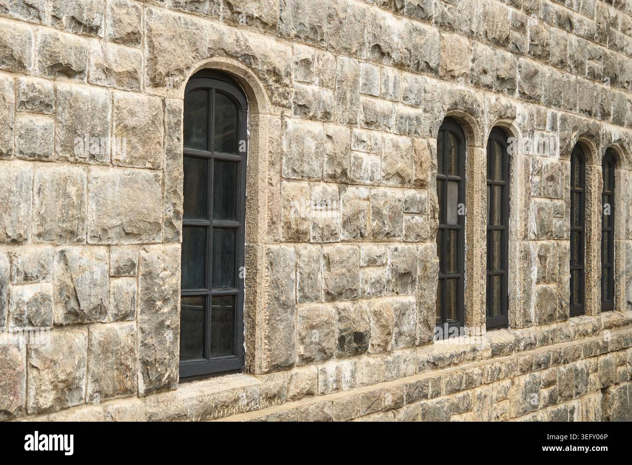 Alte schwarze bogenförmige Fenster auf langer grauer Steinmauer, Gebäudefassade mit ungleichmäßigen gemeißelten Blöcken, Seitenblick, altes Architekturkonzept Stockfoto