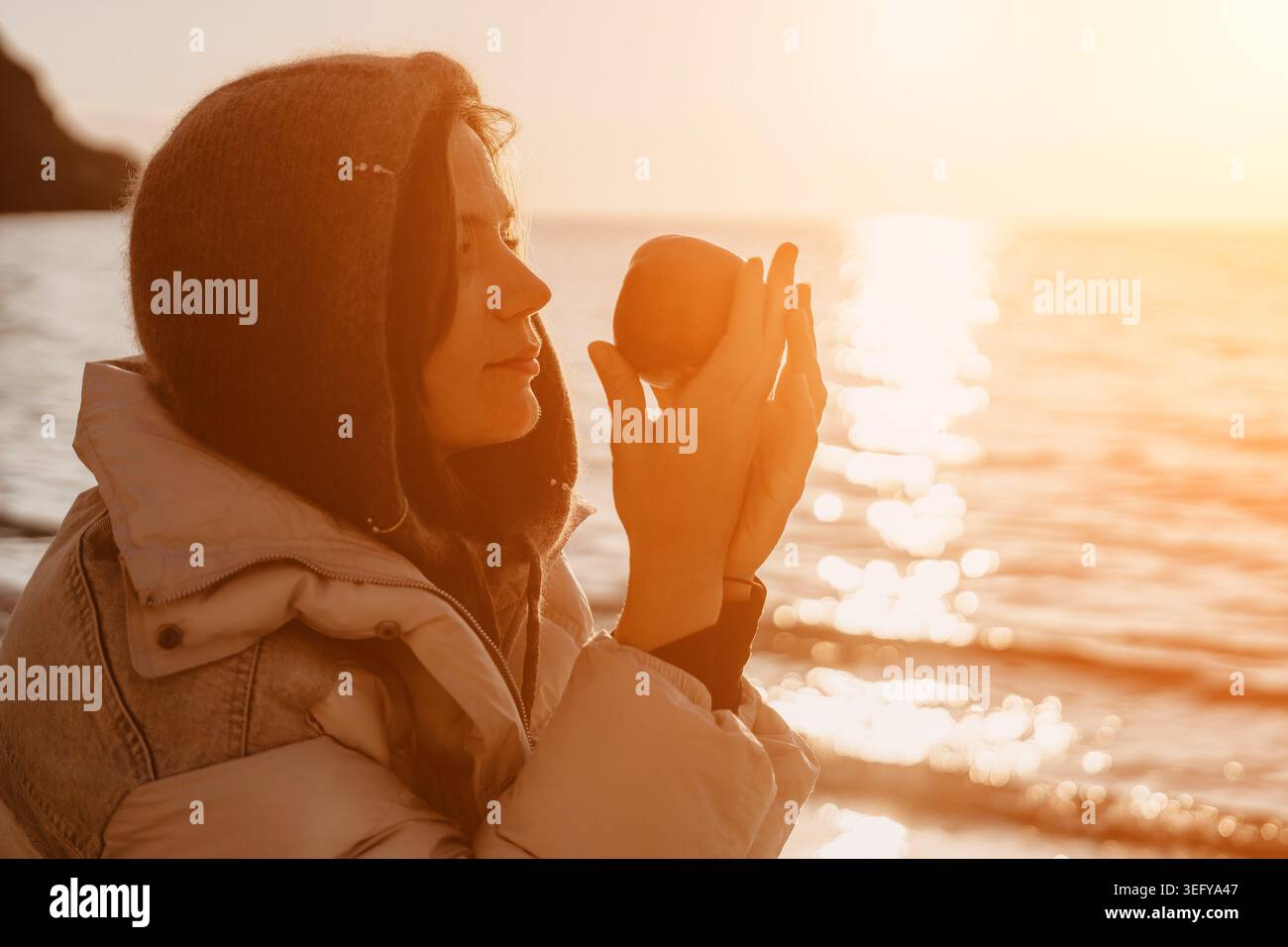 Frau Apfel Sonnenuntergang ruhige junge Frau, die einen warmen Hoodie trägt, hält einen frischen Apfel am Strand während der goldenen Stunde Stockfoto