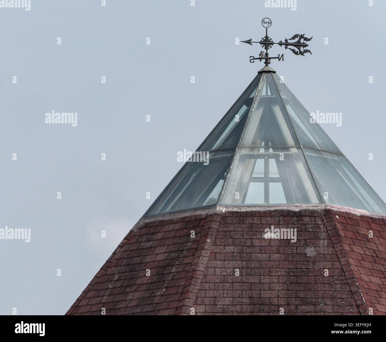 Aus nächster Nähe ein spitzes Gebäudedach mit einer Glaspyramidenstruktur, gekrönt von einer dekorativen Metallwetterfahne vor einem klaren, hellen Himmel Stockfoto