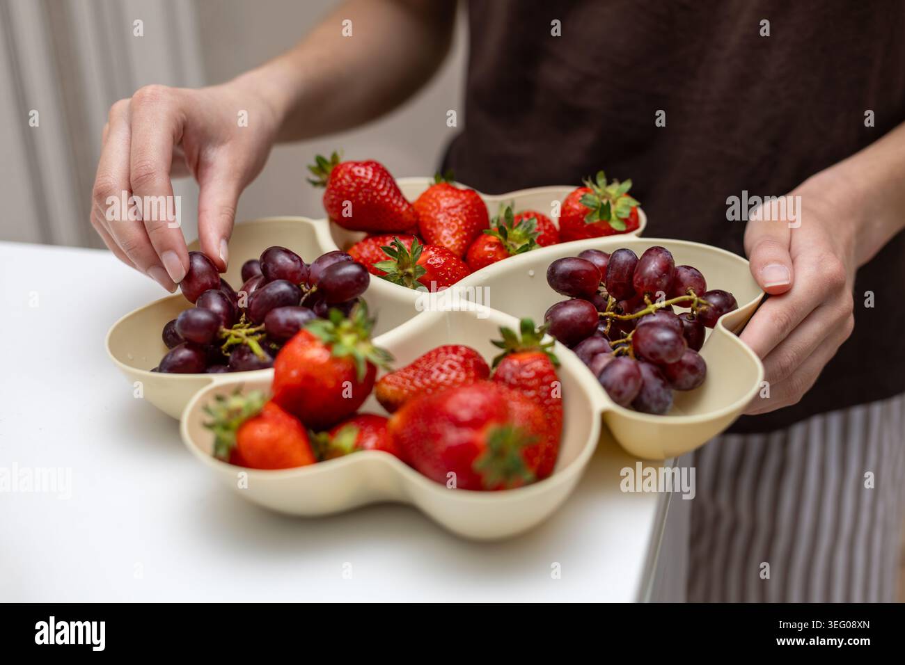 Trauben Von Hand Aus Dem Frischobst-Tablett Ernten. Nahaufnahme einer Person, die rote Trauben aus einem geteilten Tablett mit Reifen Erdbeeren für ein gesundes wählt Stockfoto