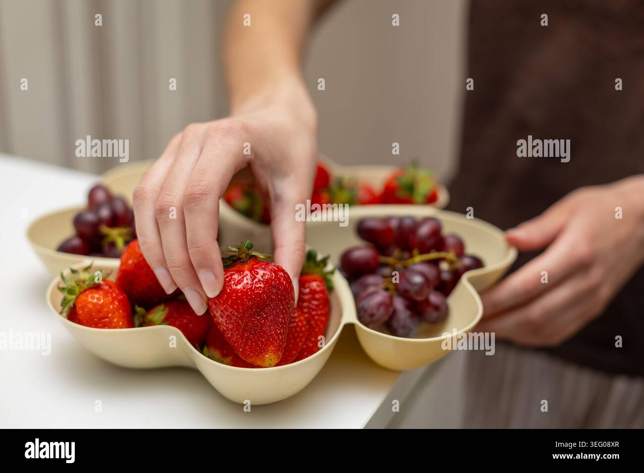 Frische Erdbeeren Aus Der Obstschale Von Hand Auswählen. Nahaufnahme einer Person, die reife Erdbeeren aus einem geteilten Obsttablett mit roten Trauben für auswählt Stockfoto