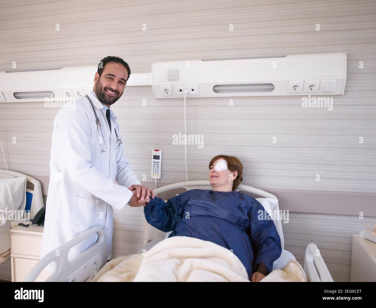 Ein männlicher Arzt aus dem Nahen Osten schüttelt mit einer sich erholenden Patientin, die in einem modernen Krankenhauszimmer eine Augenpflaster trägt, die Hand und vermittelt Vertrauen, mitfühlende Pflege und positive postoperative Erholung. Stockfoto