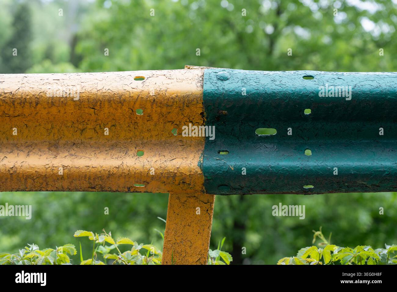 Eine verwitterte Verkehrsbarriere oder ein Sicherheitszaun aus Metall mit abblätternden Farbschichten, vor einem sanft verschwommenen Hintergrund frischer grüner Waldbäume. Clo Stockfoto