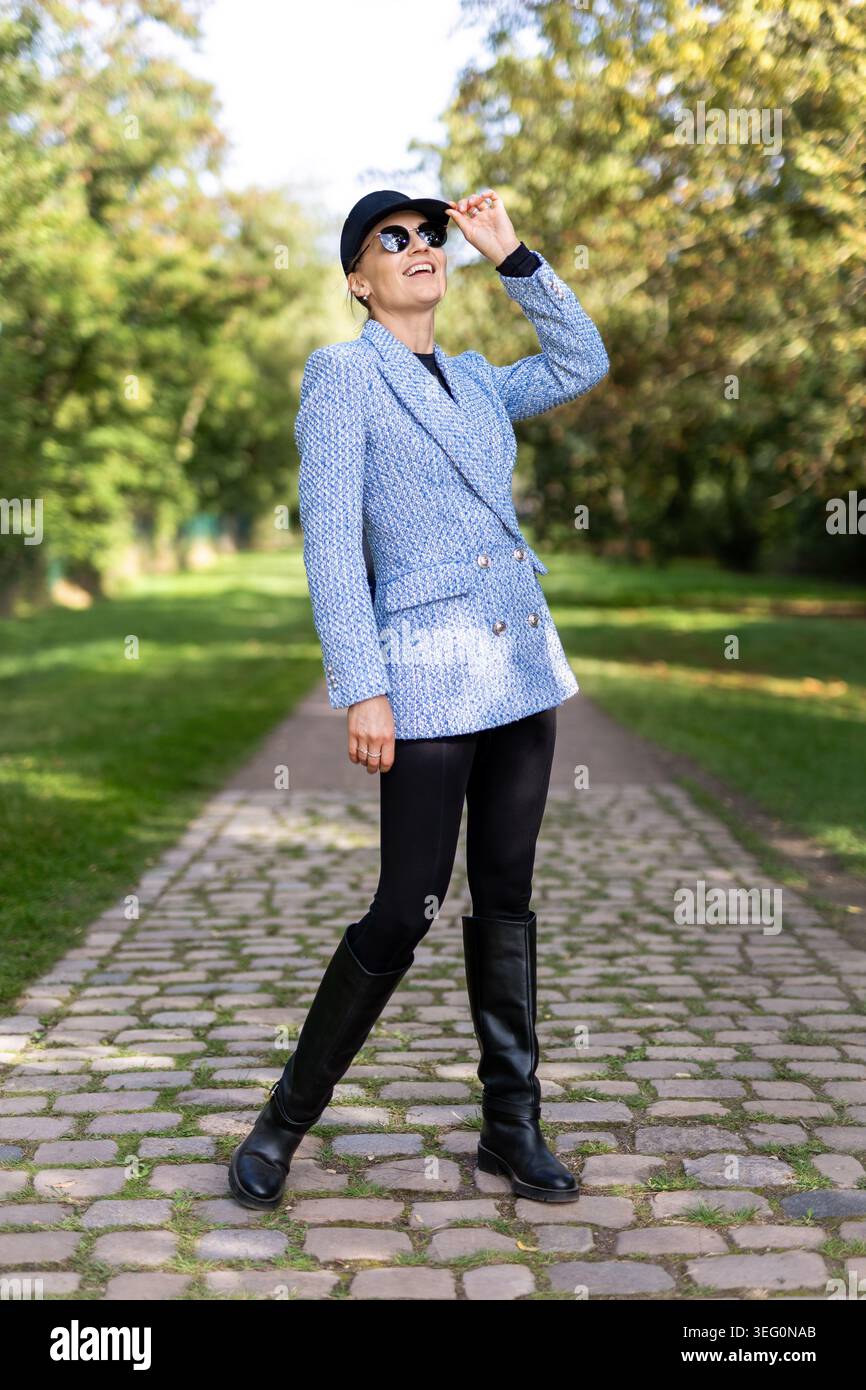 Elegante Frau, die auf dem sonnigen Parkweg spaziert, tragen Sie stilvolle Blazer Cap schwarze Stiefel. Modernes Porträt des urbanen Lifestyles mit modischem Streetstyle Stockfoto