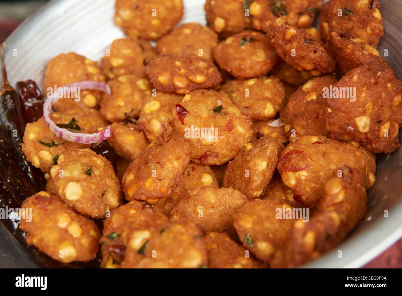 Silberne Schüssel voller knuspriger, frittierter Linsensaft Vadai-Snacks, Krapfen mit Erbsen-Chiliflocken und roher roter roter Zwiebelscheibe, Top-down-Nahaufnahme Stockfoto
