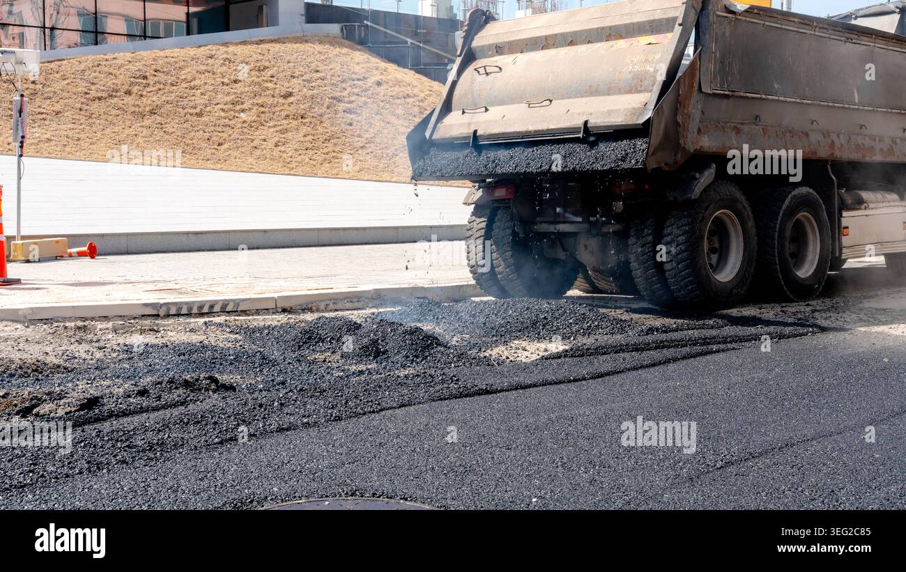 Die Arbeiter führen Asphaltpflaster auf einer städtischen Straße durch und zeigen dabei Techniken zur Straßenreparatur. Ein Lkw entlädt frischen Asphalt und hebt die Entwicklung der Infrastruktur hervor Stockfoto