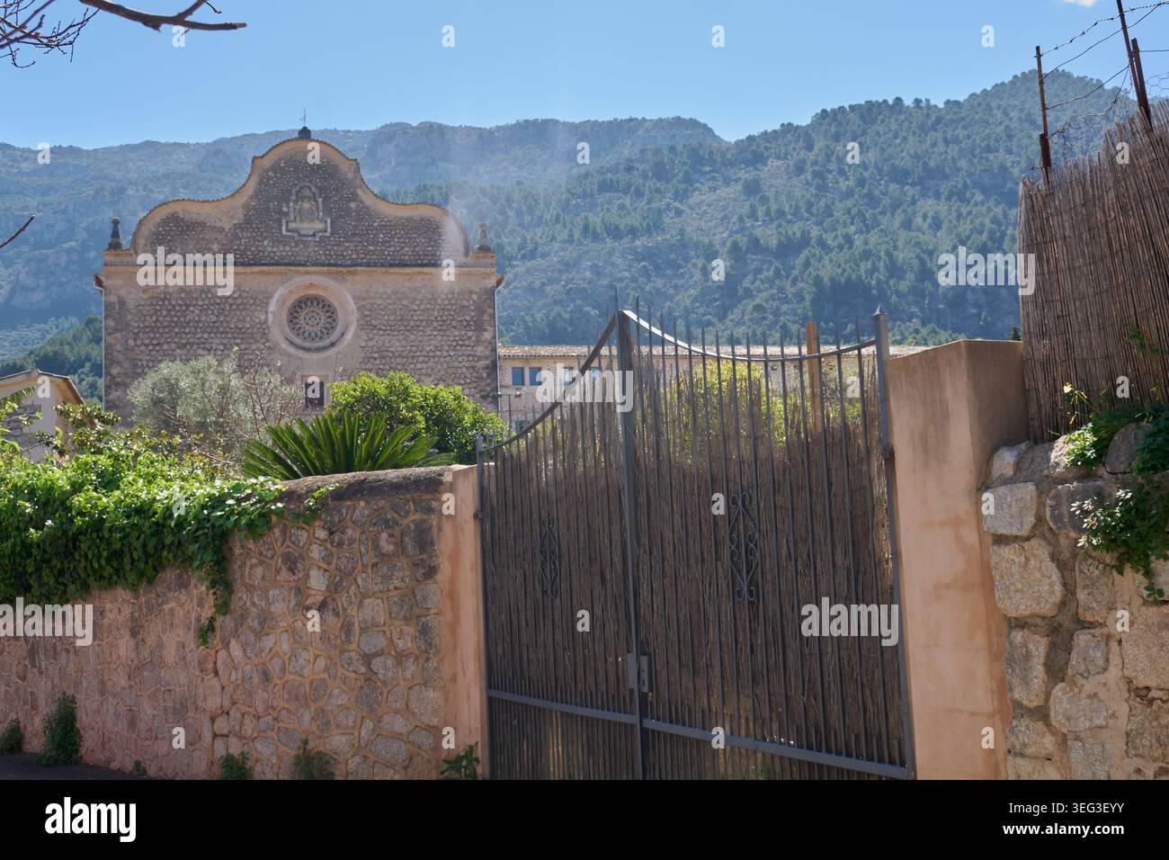 Historische Steinkirchfassade mit unverwechselbarer barocker Silhouette und Rosenfenster hinter einem rustikalen Eisentor auf dem Tramuntana-Berg Stockfoto