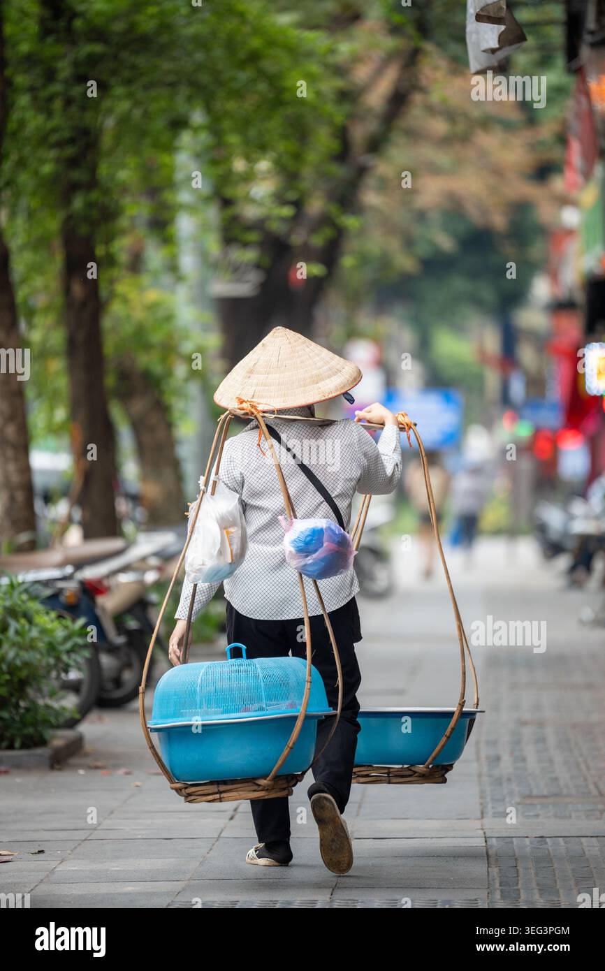 Nicht erkennbarer Straßenverkäufer mit konischem Hut, der in Hanoi die Straße hinunter läuft und Sachen auf der Schulterstange trägt. Stockfoto