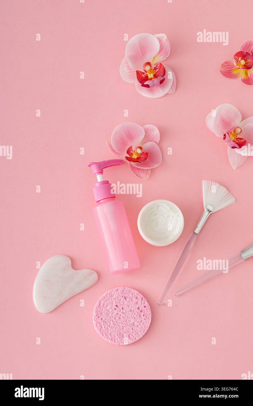 Trendige Selbstpflege- und Hautpflegekomposition mit Gesichtscreme, Kosmetikflasche, Schönheitszubehör und Orchideenblüten auf pastellrosa Hintergrund. Stockfoto