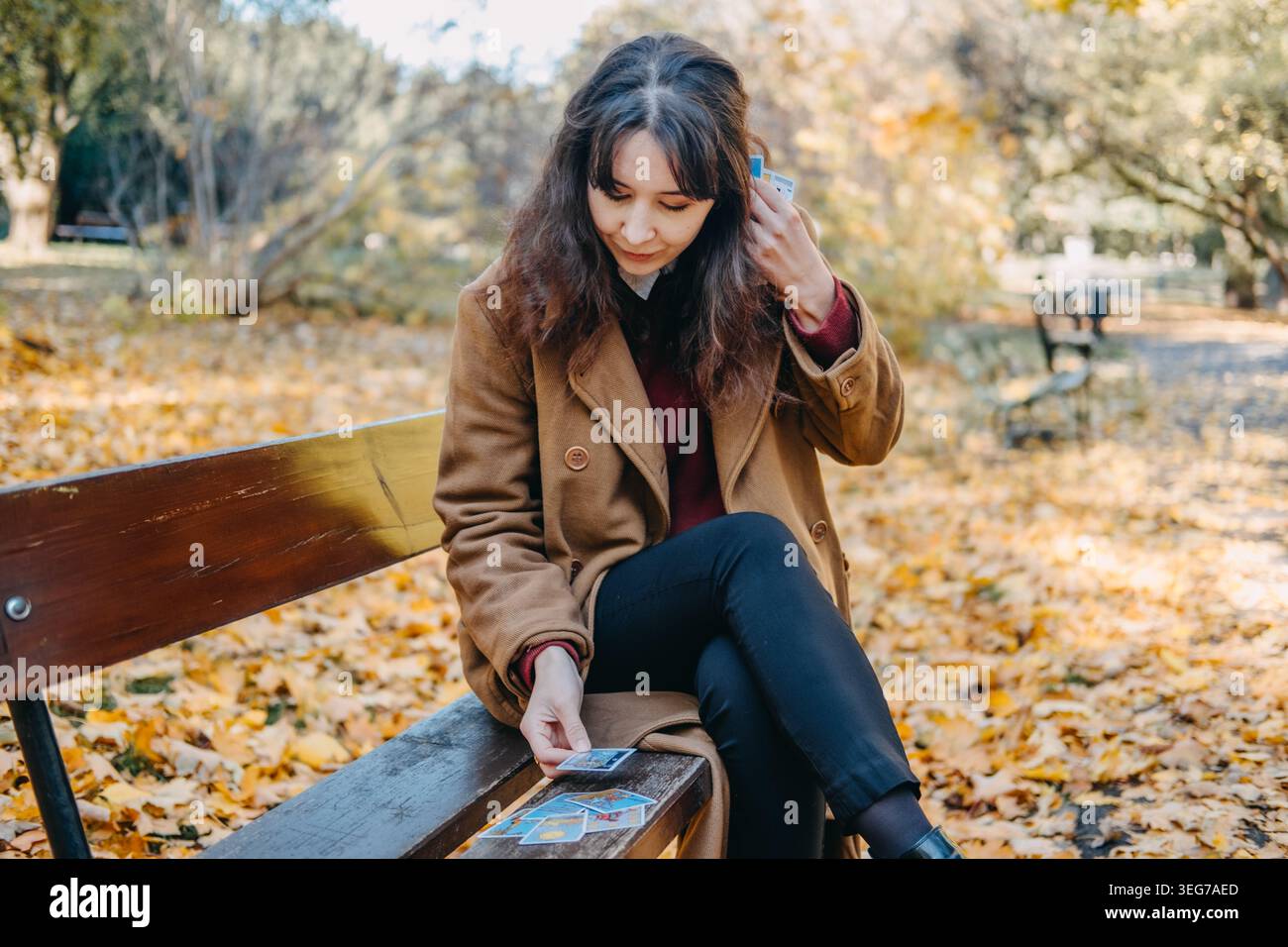 Die Frau sitzt auf einer Holzbank im Herbstpark und hält eine Tarotkarte in der Nähe ihres Gesichts. Glückssagen im Freien, Weissagungswerkzeuge, Hexerei üben Stockfoto
