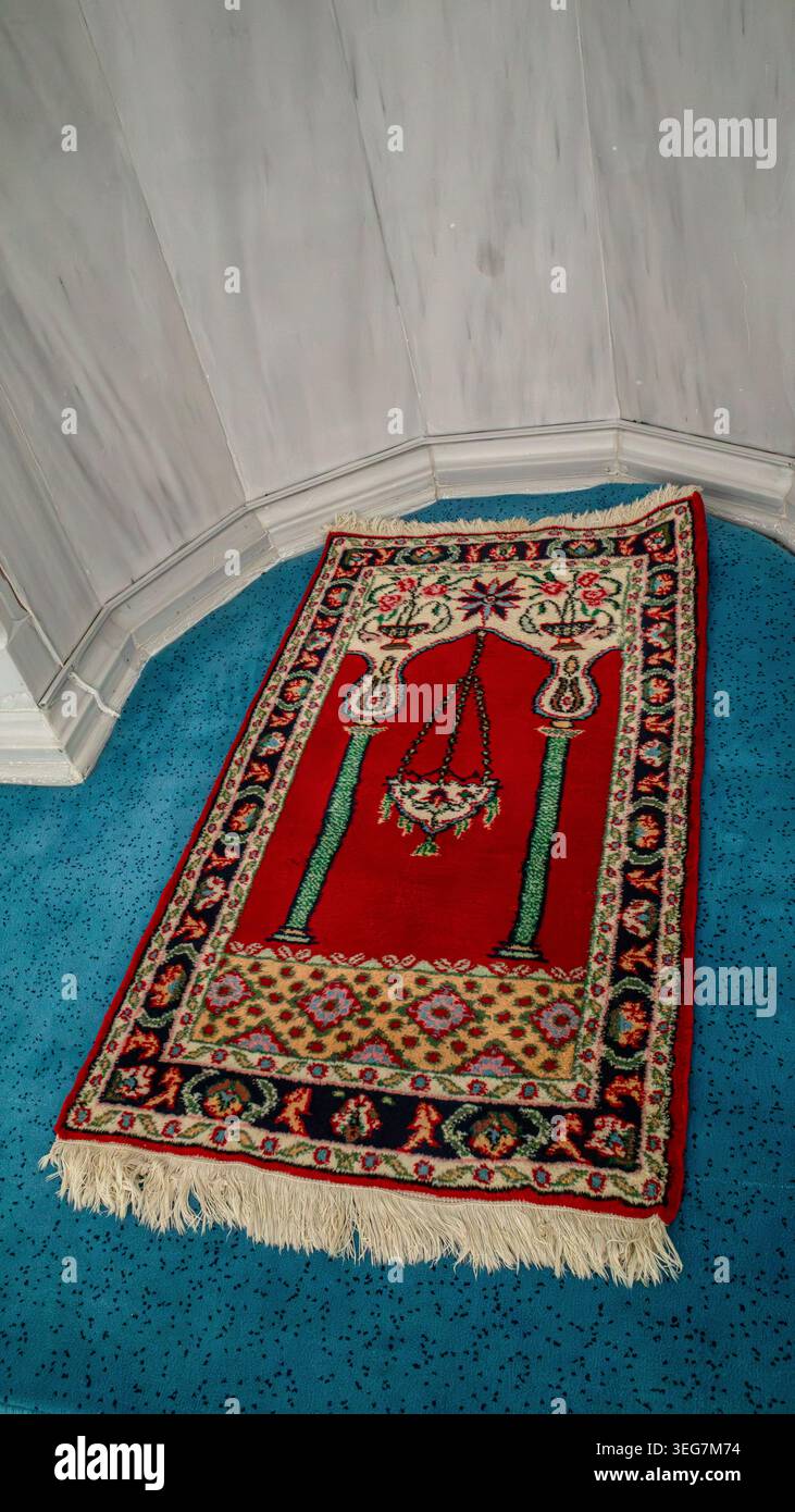 Das Foto zeigt einen roten Gebetsteg auf einem blauen Teppichboden in einem modernen Gebetssaal. Der Teppich ist mit Blumenmotiven und Moscheesymbolen versehen. Stockfoto