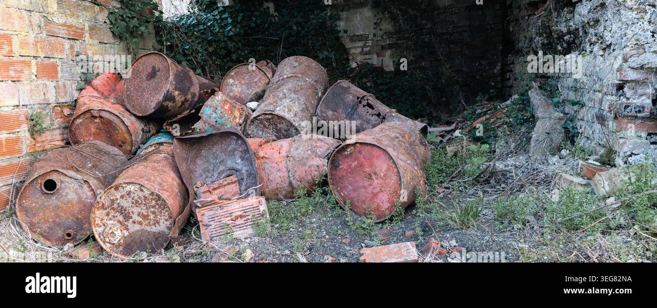 Ein Haufen rostiger Metallfässer und zerquetschter Trommeln sitzt in einem verwachsenen, verlassenen Schuppen, umgeben von Efeu, gebrochenen Ziegeln und Unkraut, was an Verfall und Negle erinnert Stockfoto