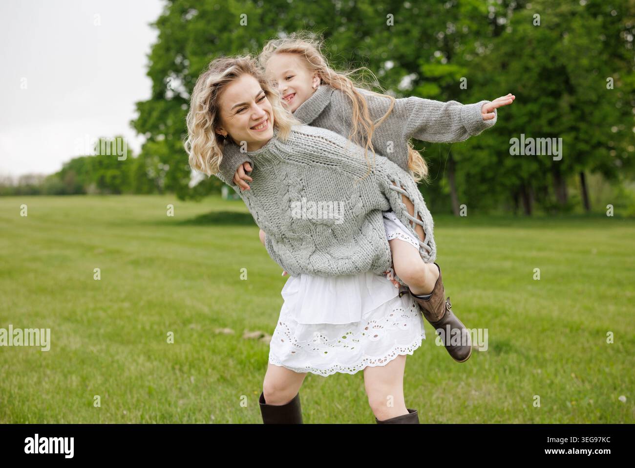 Eine fröhliche Mutter trägt ihre Tochter auf dem Rücken in einem üppigen grünen Feld und zeigt ihre Verbundenheit und ihr Glück in einer verspielten Outdoor-Umgebung. Stockfoto
