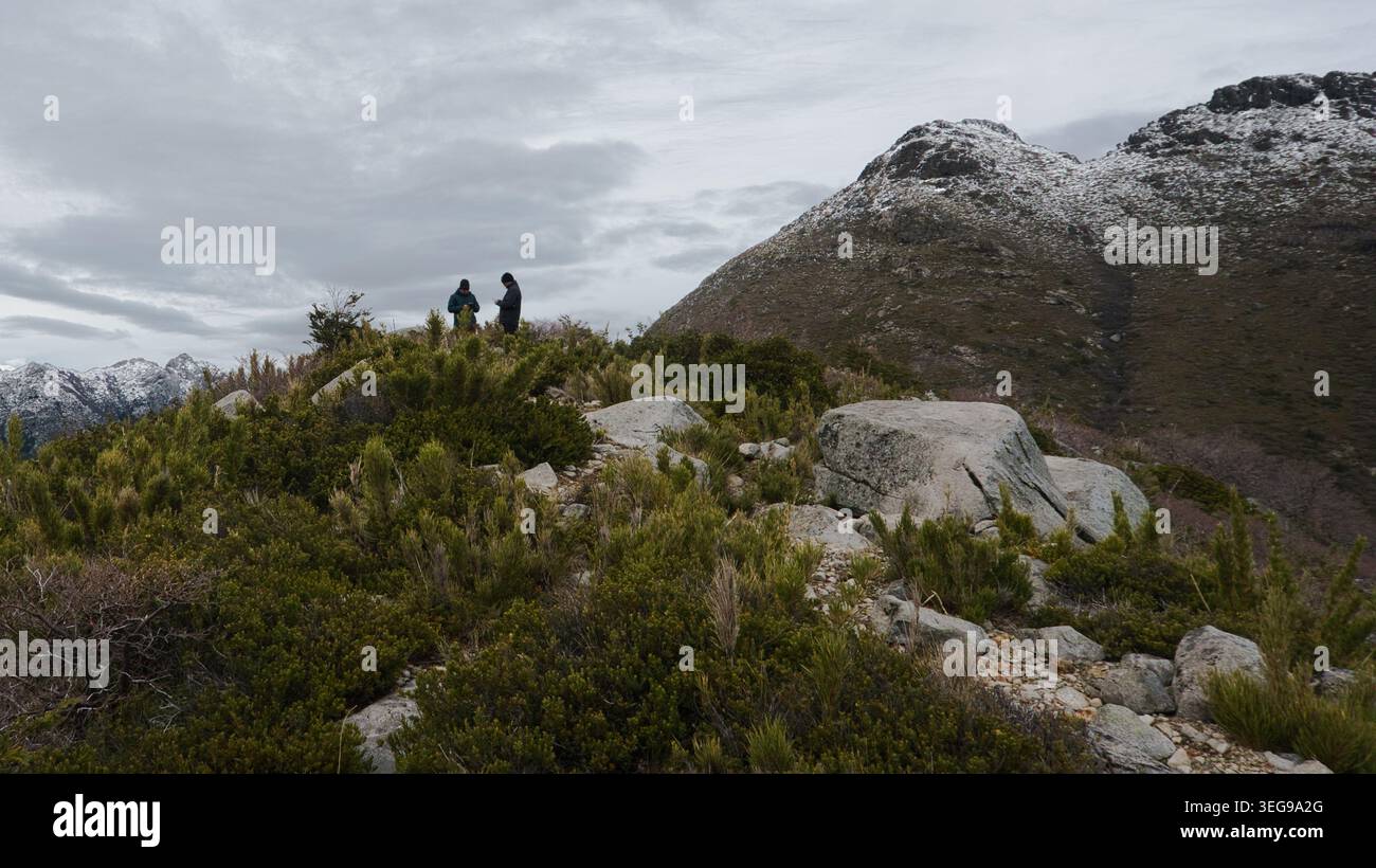 Zwei Wanderer, die auf einem zerklüfteten Bergkamm stehen und die Aussicht mit schneebedeckten Gipfeln unter einem dramatischen bewölkten Himmel in den südlichen Anden Chiles genießen. Stockfoto