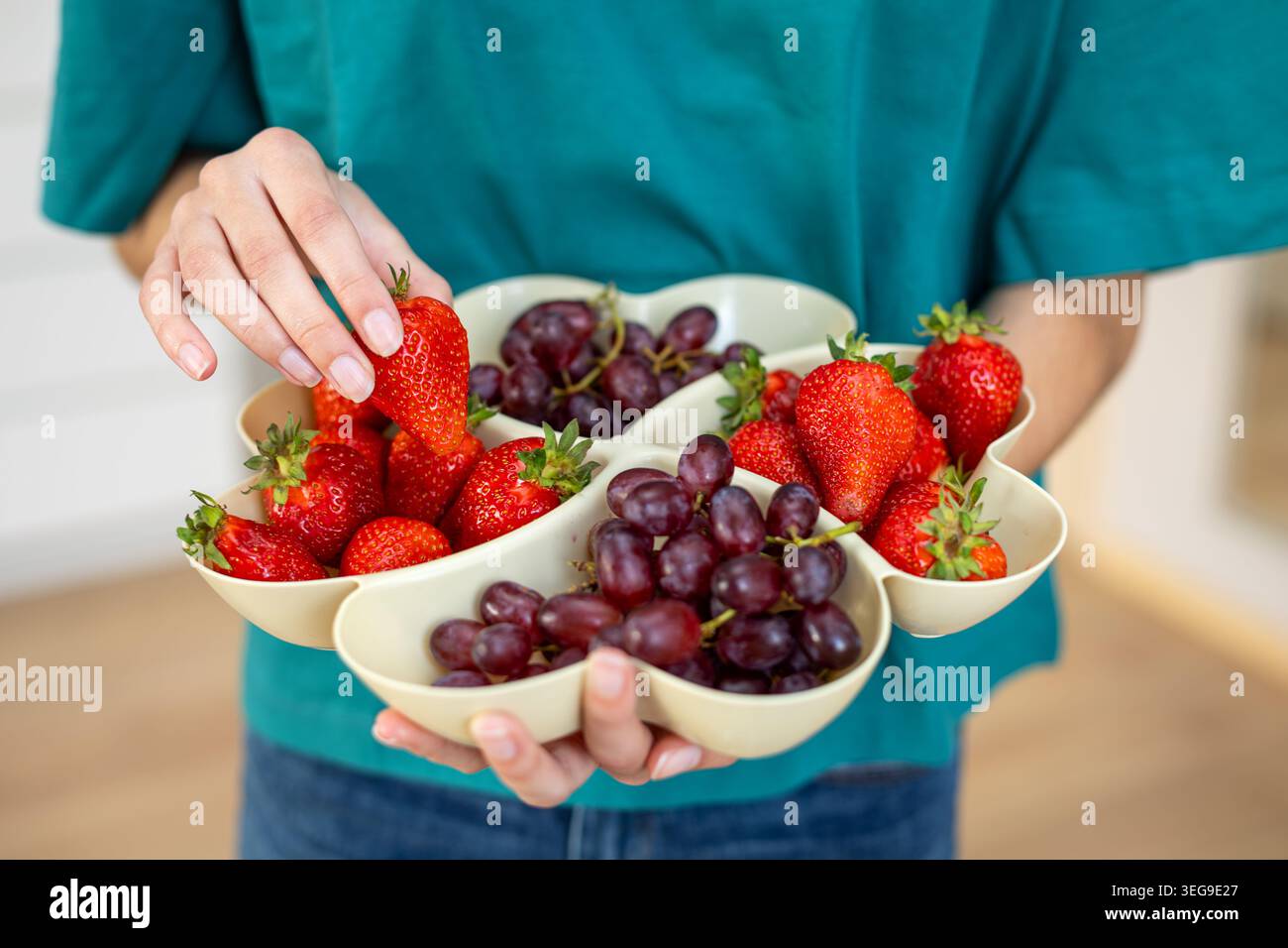 Frische Erdbeeren und Trauben auf Servierplatte. Person, die ein geteiltes Obsttablett mit frischen Erdbeeren und roten Trauben hält, perfekt für gesunde Stockfoto