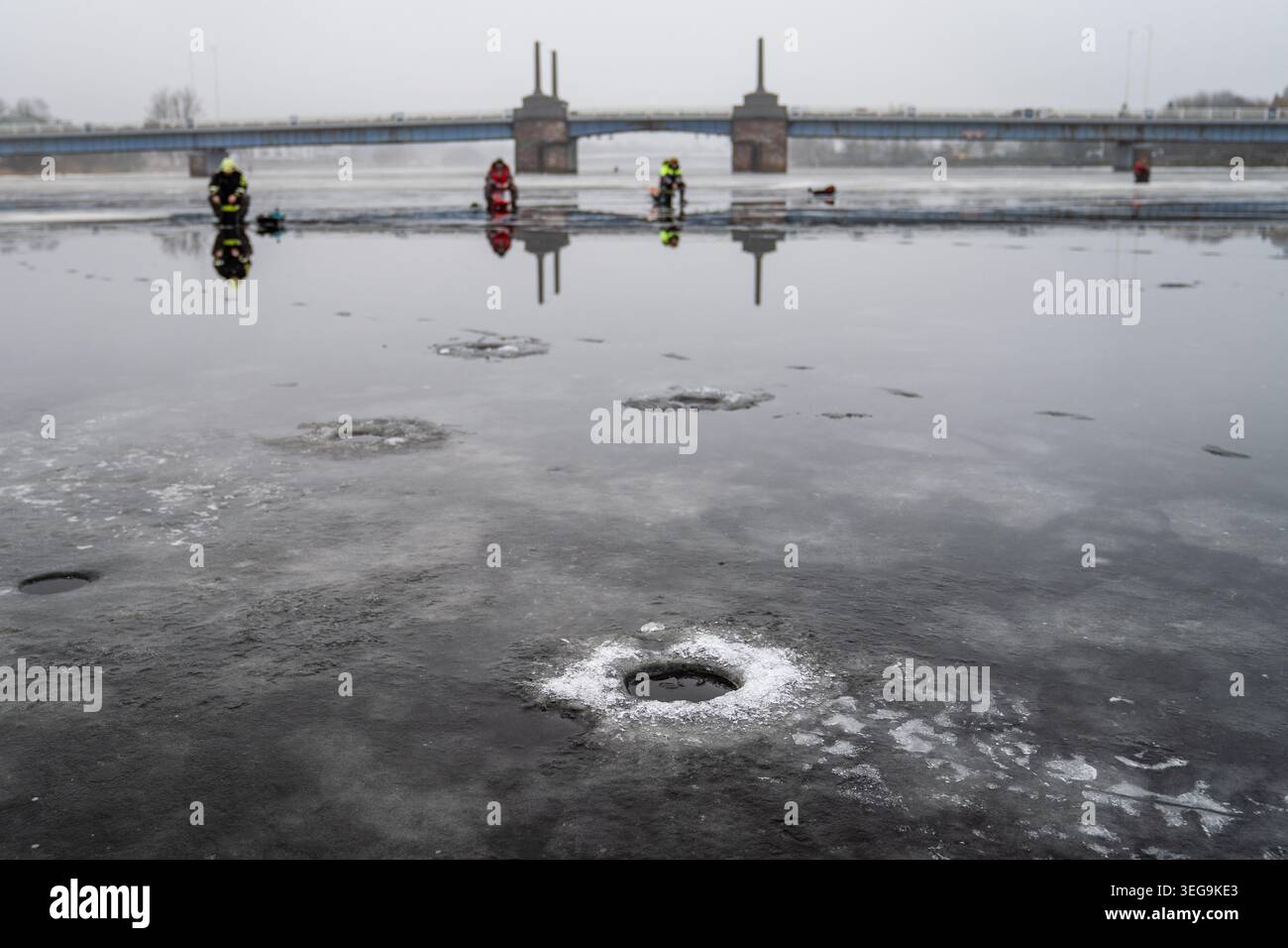 Das dunkle Eis des Pärnu-Flusses ist durch eine Reihe von Angellöchern geprägt. In der Ferne sitzen Fischer bewegungslos vor der Silhouette der Kesklinna Bridg Stockfoto