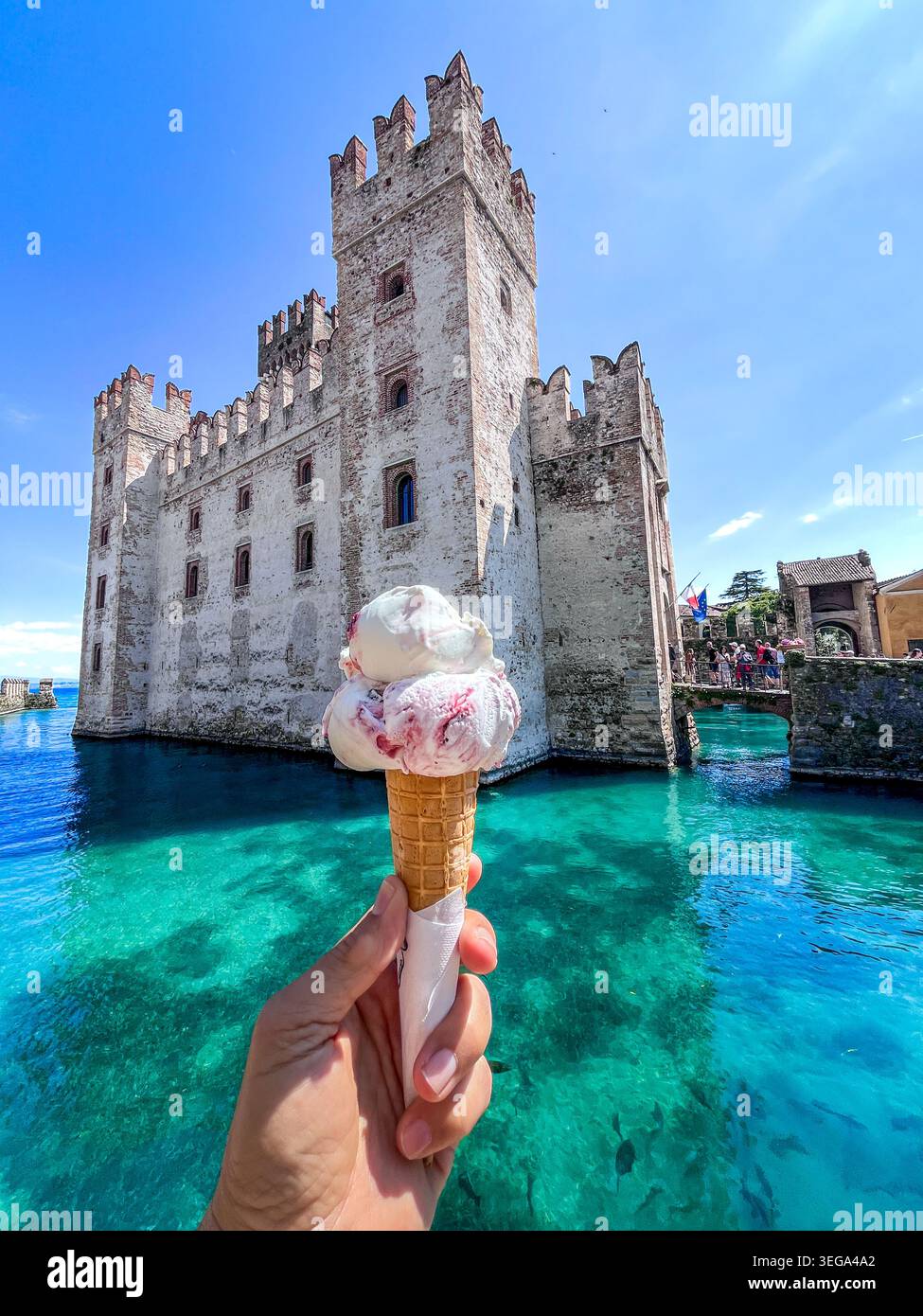 Eine Hand hält einen köstlichen Eiskegel mit doppelter Schaufel vor dem historischen Schloss Scaligero, umgeben vom türkisfarbenen Wasser des Gardasees in S - Smartphone-aufgenommenes Stockfoto