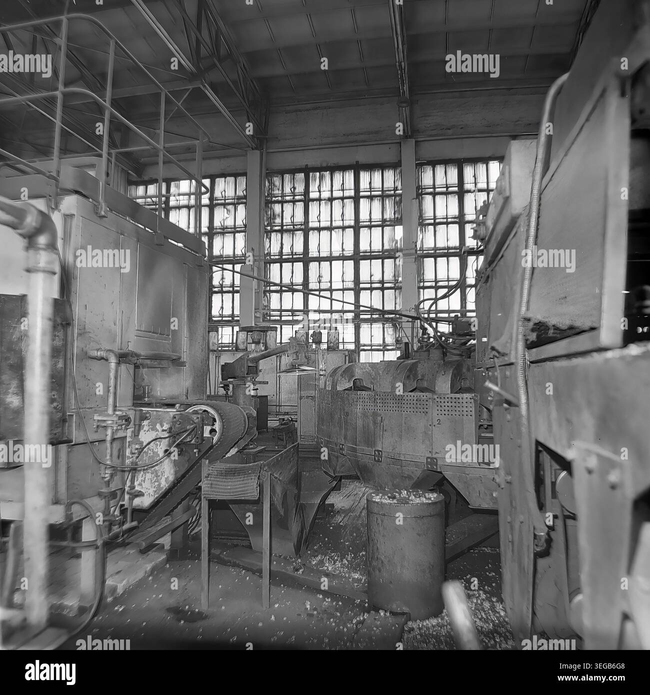 Sloviansk, 1981, AIZ (Armature-Insulator Plant). Eine leere Ansicht der Fabrikhalle mit schweren Industriemaschinen, einem Fördermechanismus und einem Metallbehälter, der mit entsorgtem Material gefüllt ist. Sonnenlicht strömt durch eine massive Wand aus mehrverglasten Industriefenstern im Hintergrund und beleuchtet den Arbeitsbereich. Ein historisches Schwarzweiß-Archivfoto, das die authentische Atmosphäre der Epoche, die Größe der Produktionsanlagen und die industrielle Umgebung feststellt. Ein detaillierter Einblick in die Vergangenheit, die sowjetische Industrie und den friedlichen Donbass. Stockfoto