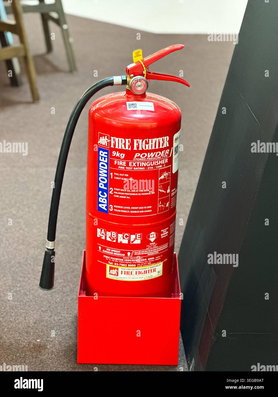 Johor, Malaysia, 13. Mai 2026: Roter 9 kg Pulverfeuerlöscher mit Schlauch und Sicherheitsstift, in Halter neben dunkler Trennwand platziert. Stockfoto