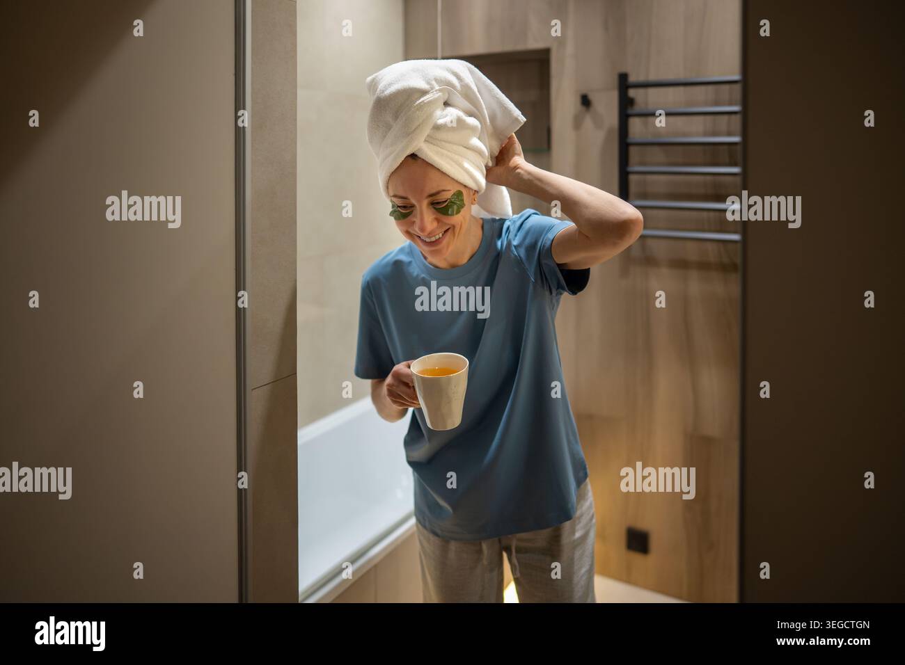 Lächelnde Frau mittleren Alters mit Handtuch auf dem Kopf, die eine Tasse Tee in moderner Badezimmereinrichtung hält. Selbstpflegeritual am Morgen oder Abend, Wellness-Lifestyle. Stockfoto