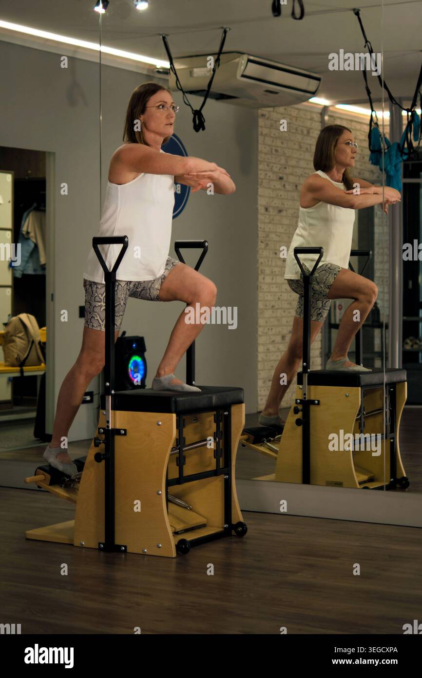 Eine fokussierte Frau übt einen kontrollierten Ausfallschritt auf einem Pilates-Stuhl in einem Spiegelstudio, betrachtet auf Augenhöhe. Die Szene spiegelt achtsame Fitness und Balance wider Stockfoto